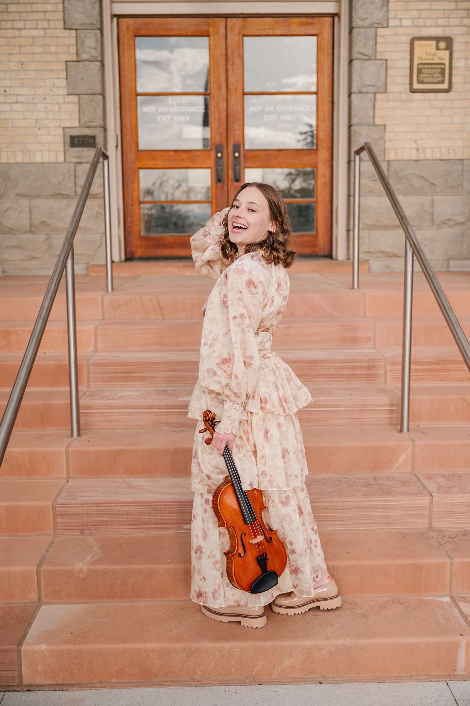 Someone in a vintage floral dress holds a violin while joyfully posing on stone steps near wooden doors.