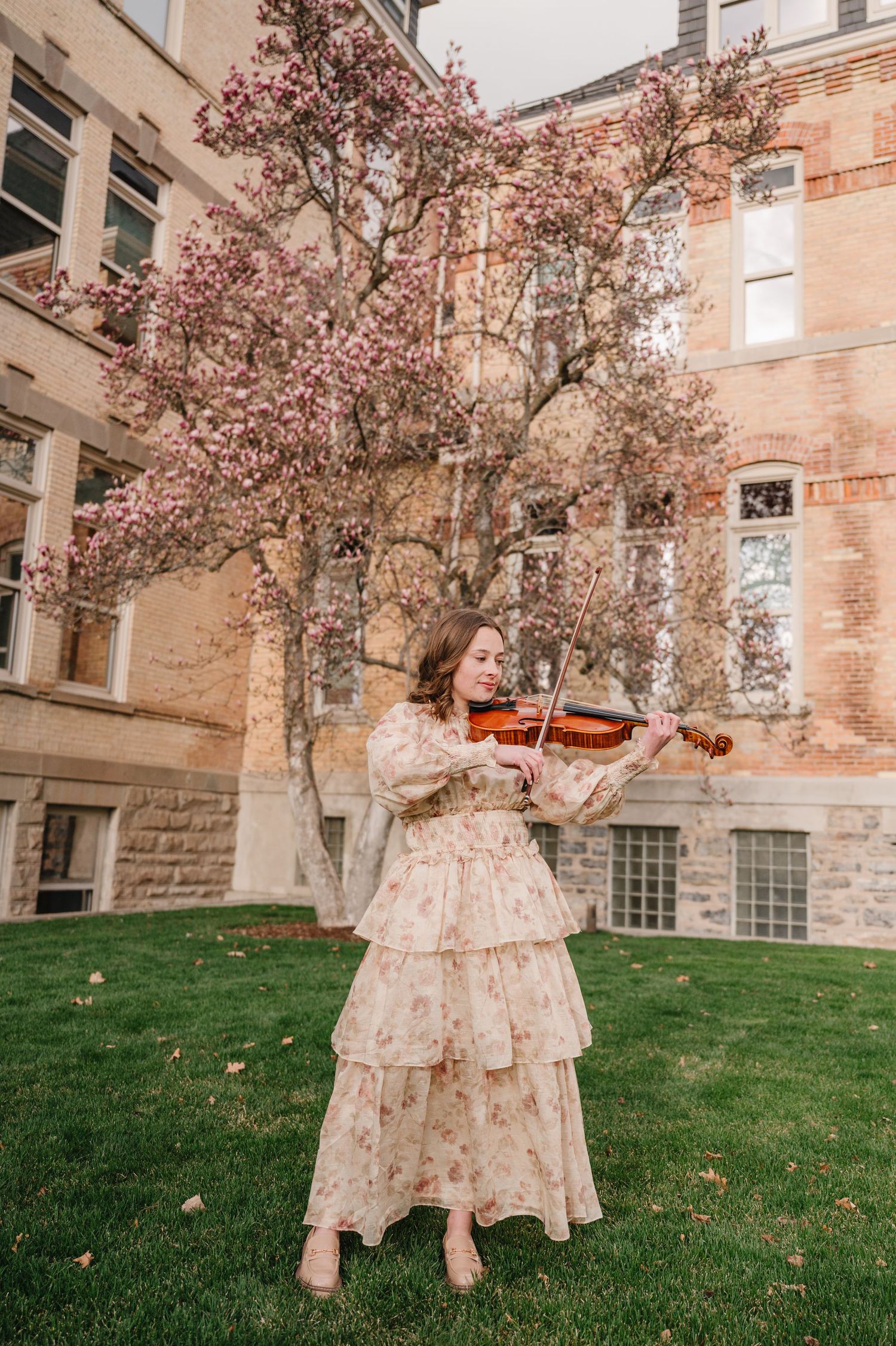 A musician in a flowing floral dress plays violin beneath pink blooming trees outside a historic stone building.