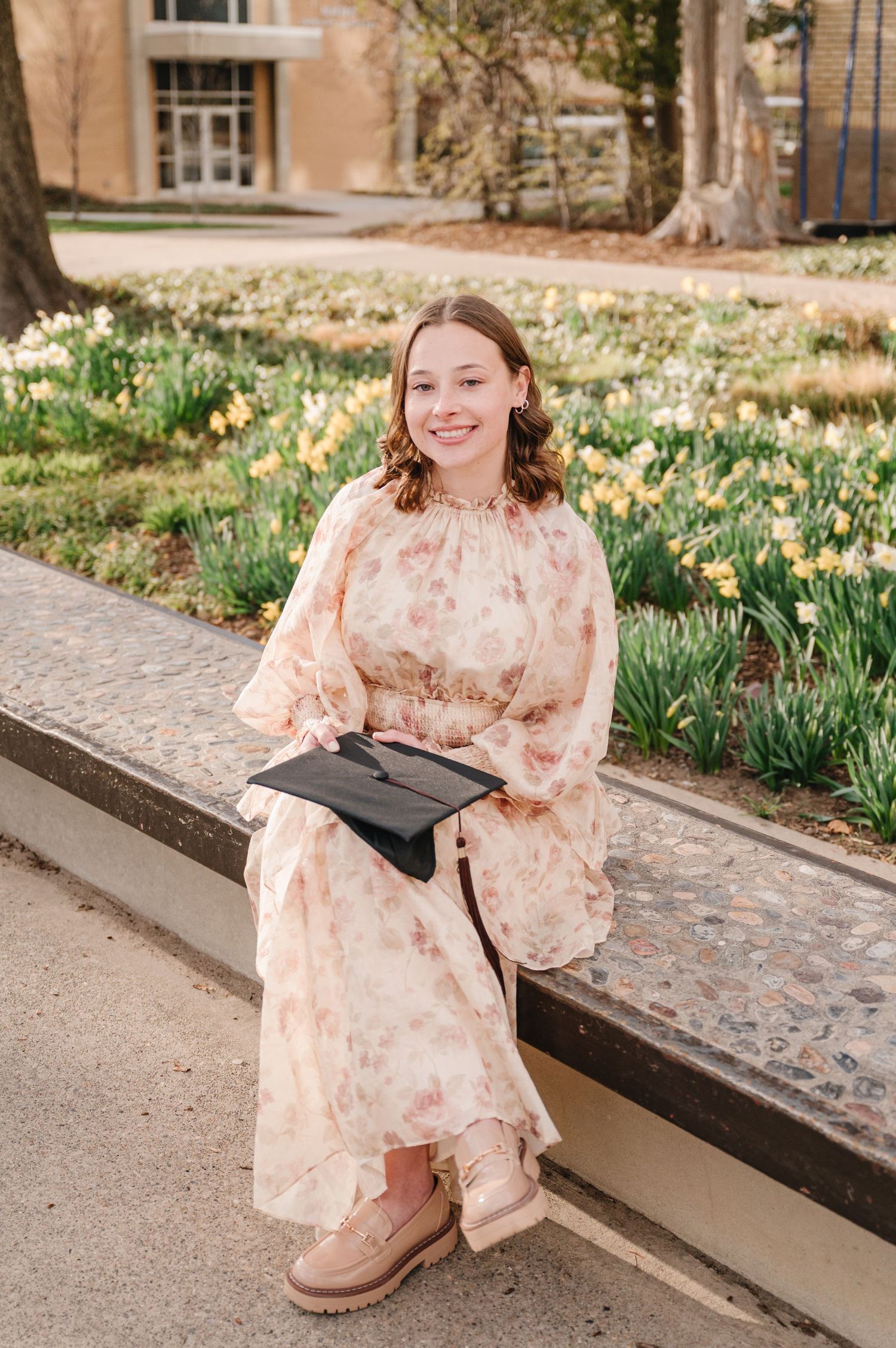 A graduate in a pale floral dress and boots sits on a stone ledge with their graduation cap amid blooming daffodils.