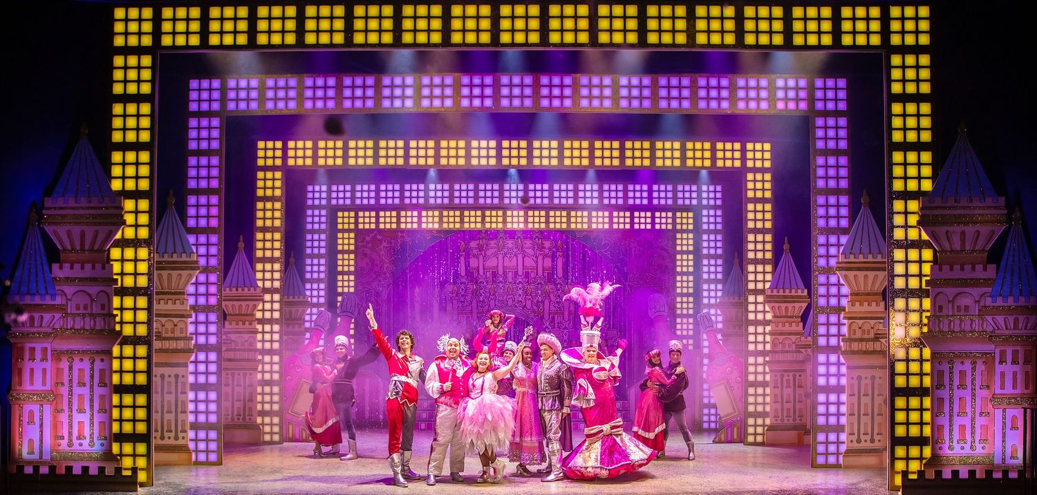 Musical theater production featuring bright yellow geometric set design and ensemble cast in colorful costumes.