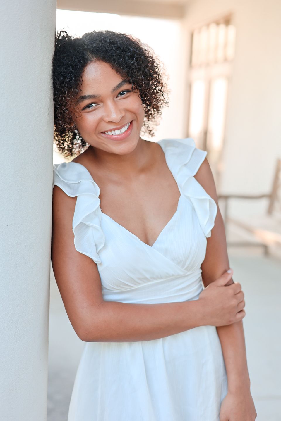 Portrait of woman in white ruffled dress smiling brightly against bright interior wall. Senior high school graduation casuals.