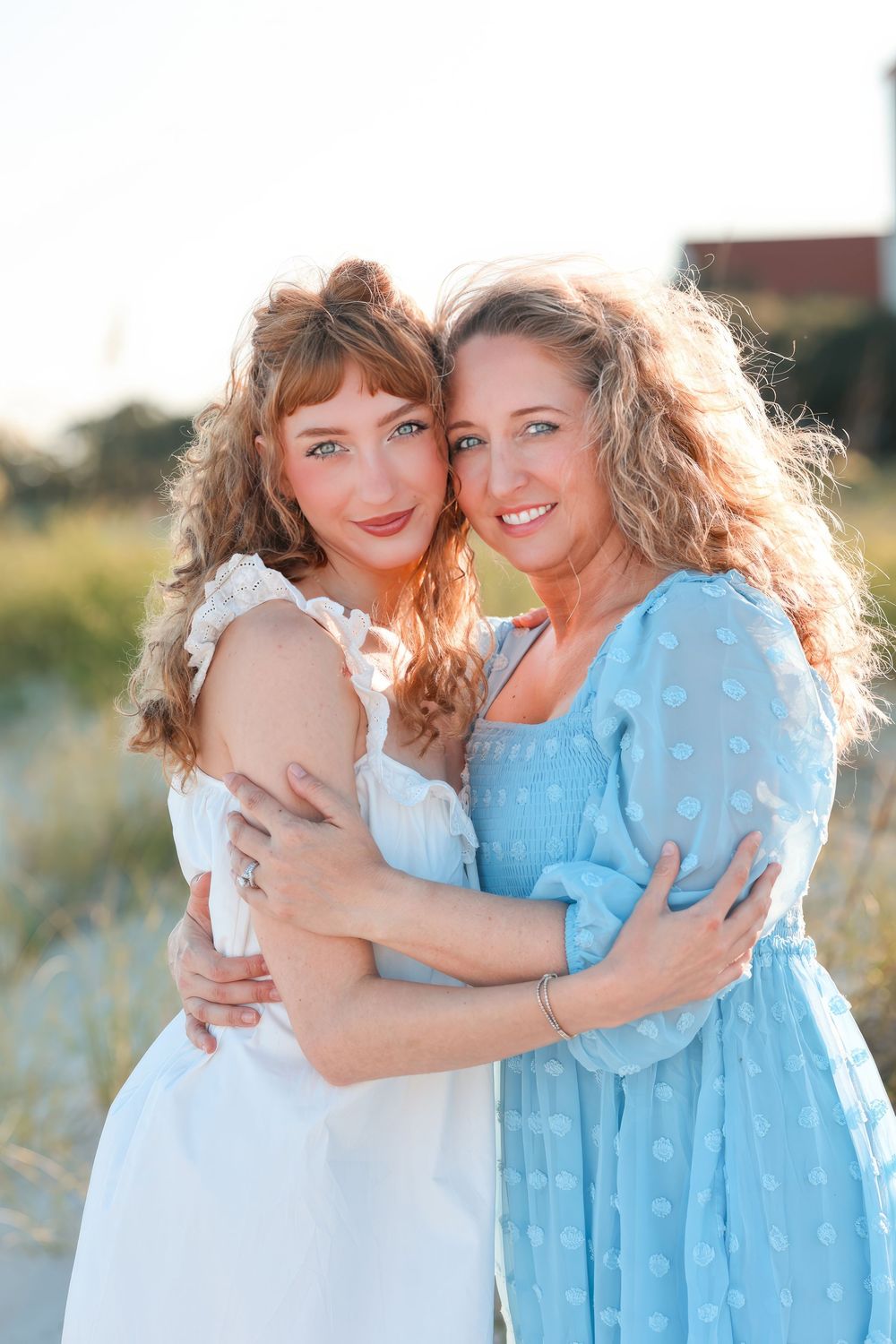 Two friends share a tender embrace outdoors while wearing white and light blue summer dresses.