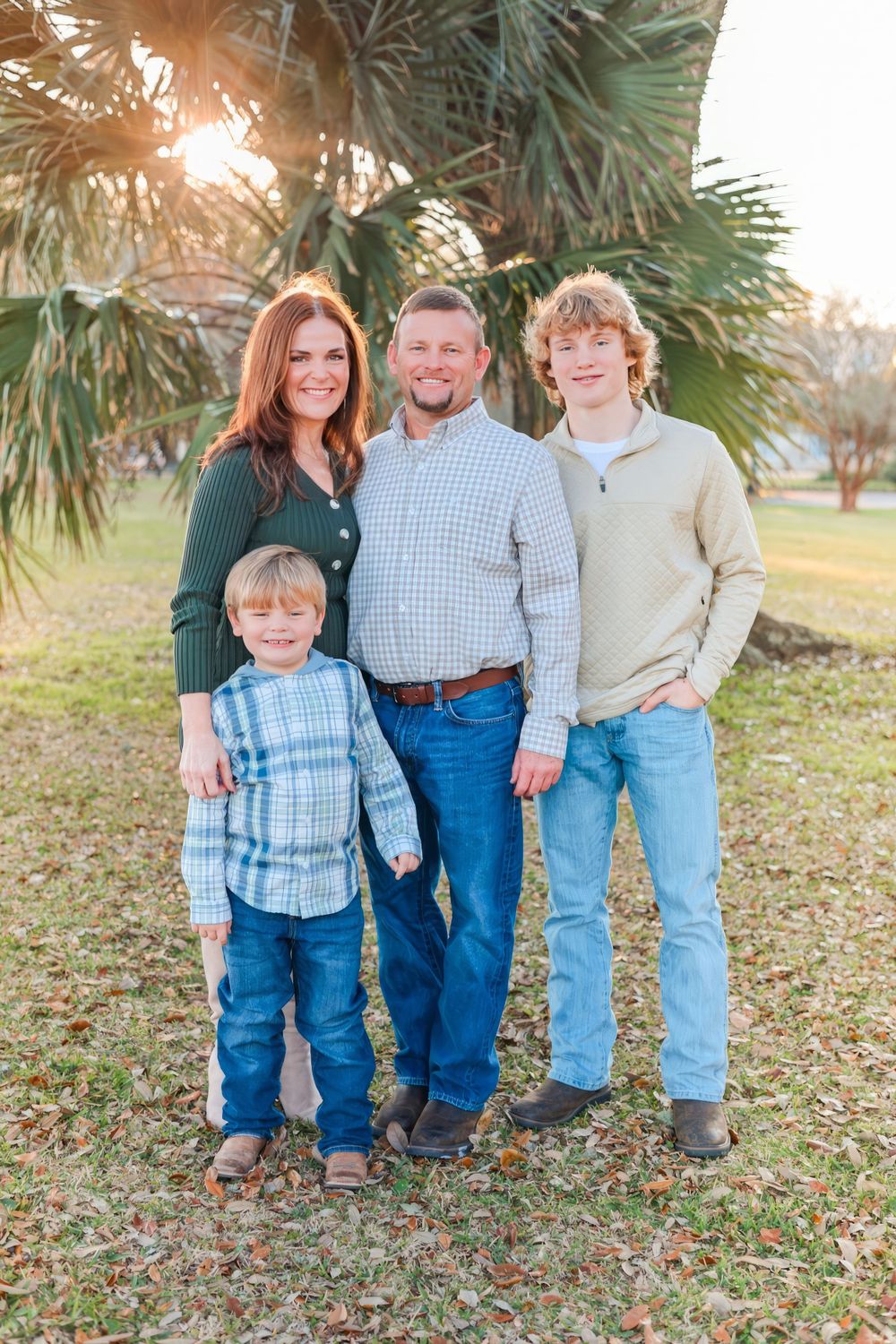 Family of four poses together outdoors with palm trees and sunset in background. Family photography session.