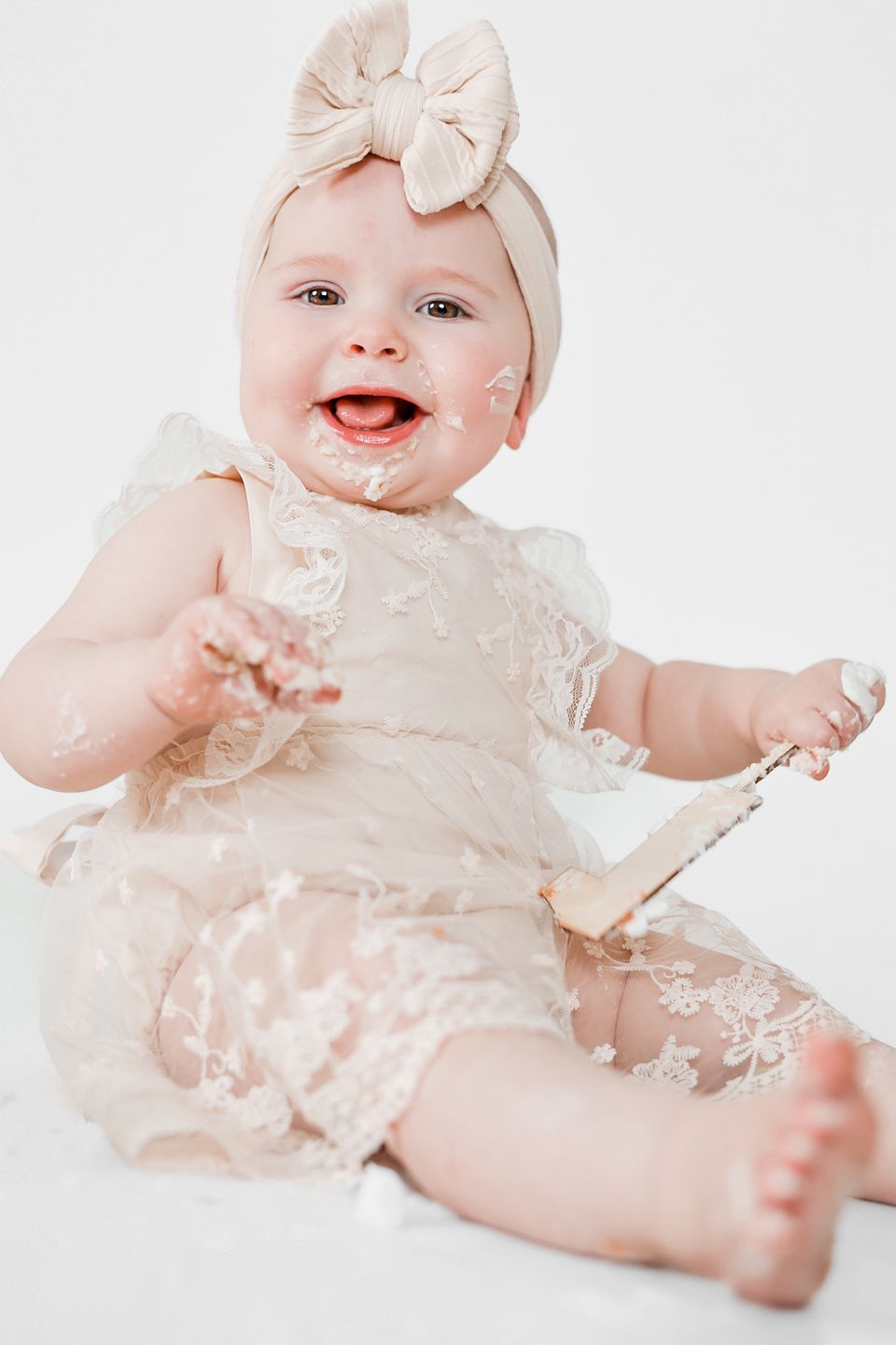 Happy baby in cream lace dress and matching bow headband sitting against white background. First birthday pictures.