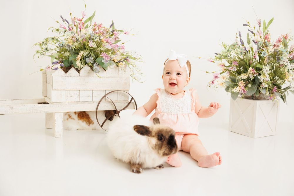 Baby girl in pink romper sits with fluffy rabbits surrounded by spring flowers in a bright white studio setting.