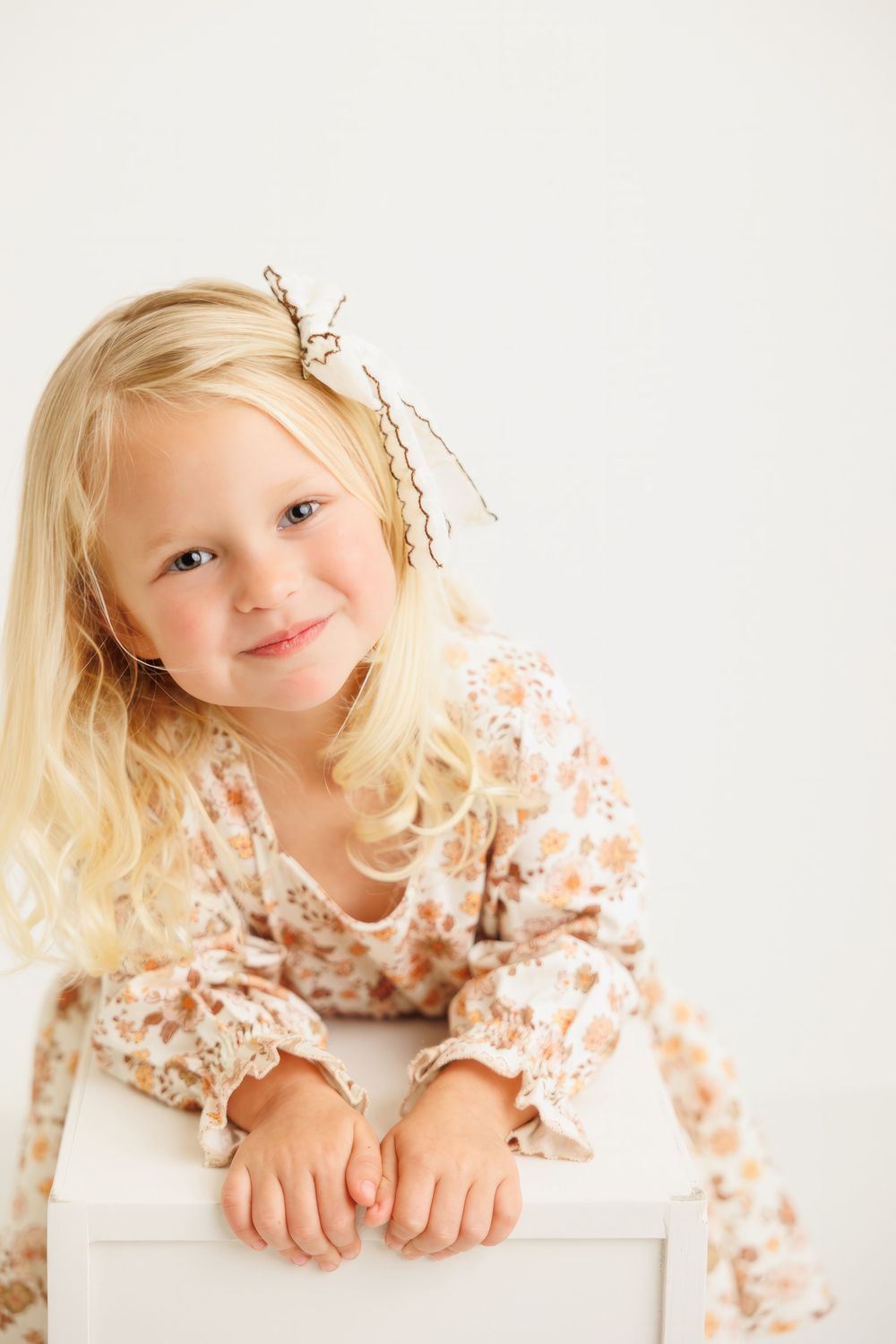 Smiling blonde toddler girl in floral dress with hair clip, leaning on white cube against light background.