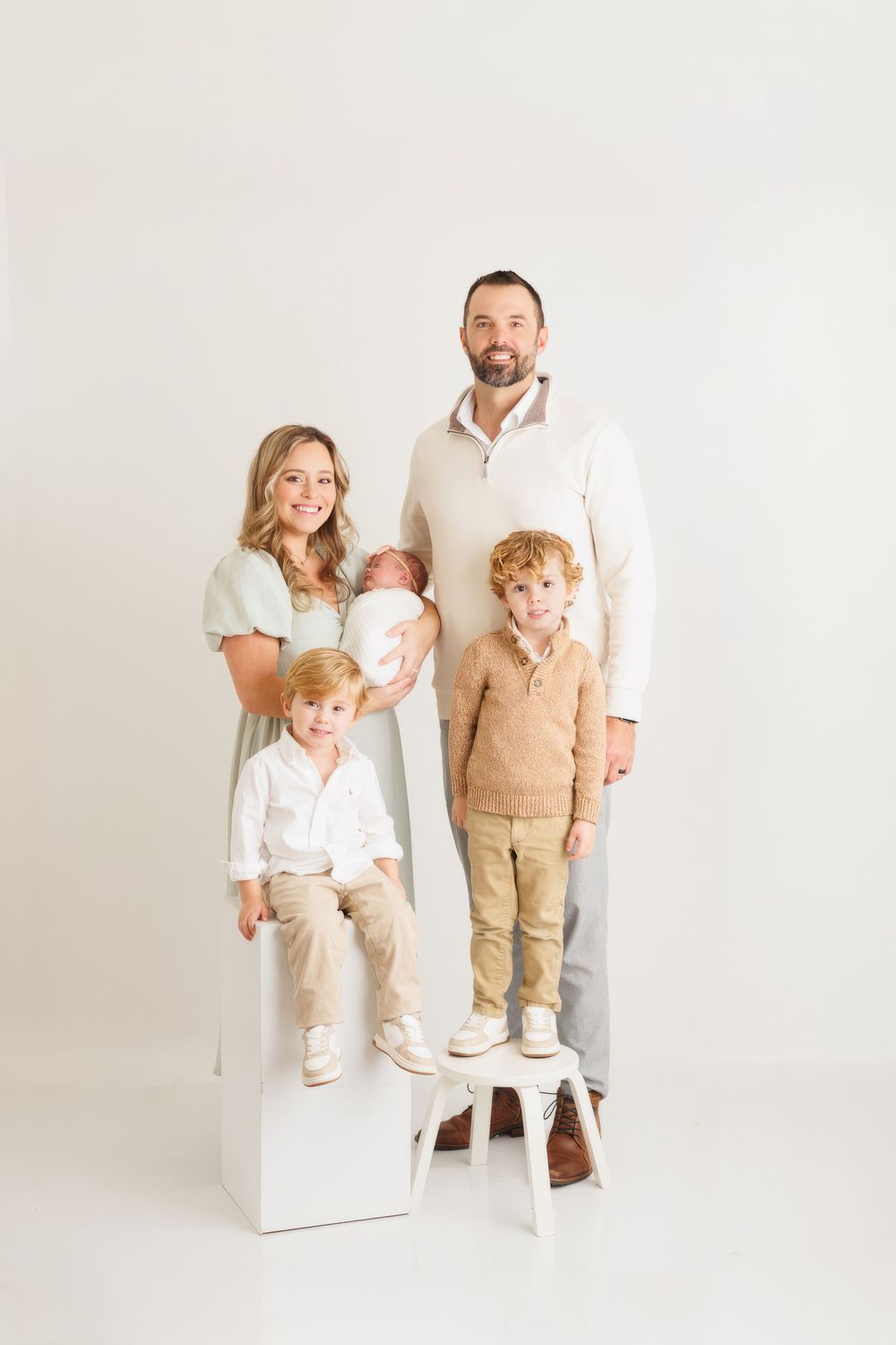 Family wearing coordinated white and beige outfits poses together in a bright studio setting for professional portraits.