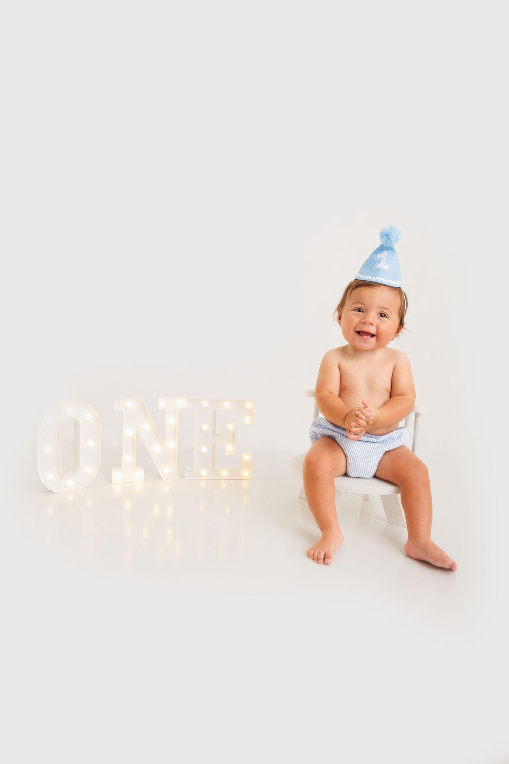 Smiling baby in blue party hat sits beside lit ONE marquee letters celebrating first birthday.