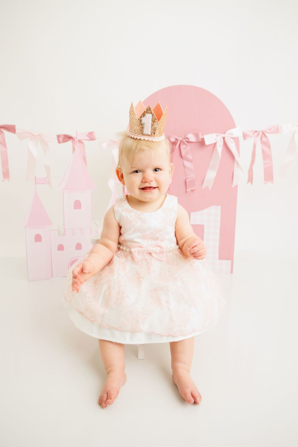 Baby girl in pink dress and crown celebrates 1st birthday with castle decorations and pink bunting banner.