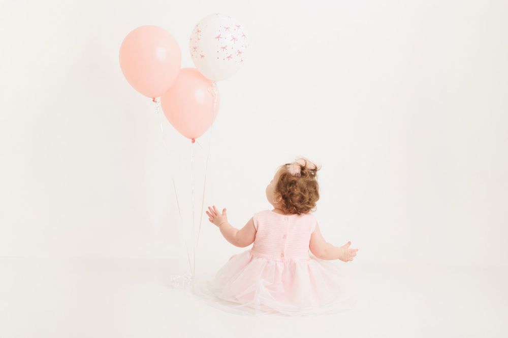 A baby in a pink dress crawls across a white floor while pink balloons float in the background.