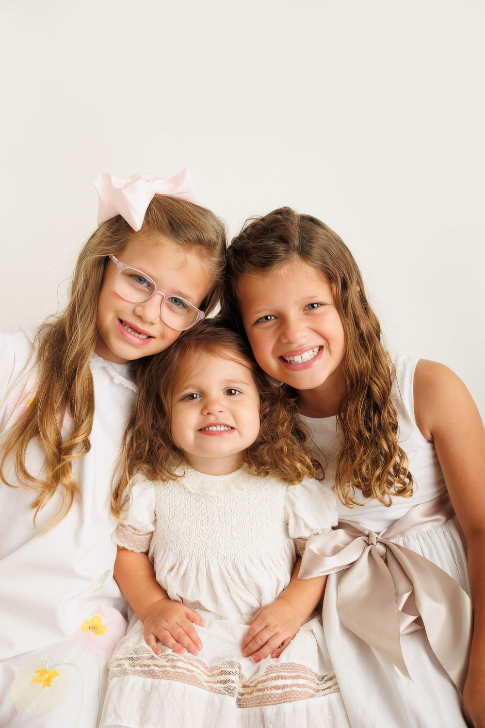 Three young sisters in white and pink dresses smiling together, oldest wearing glasses with a pink bow.