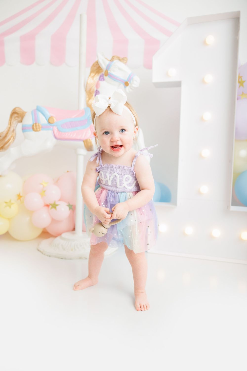 Smiling baby girl in pastel rainbow dress with bow headband at a carnival first birthday party.