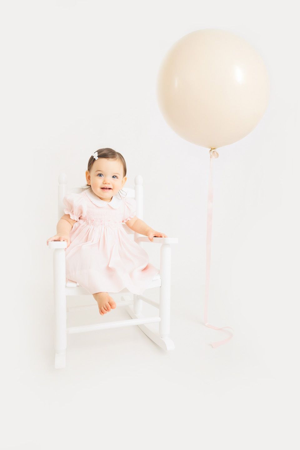 A smiling baby girl in a pink dress sits in a white rocking chair beside a large cream balloon.
