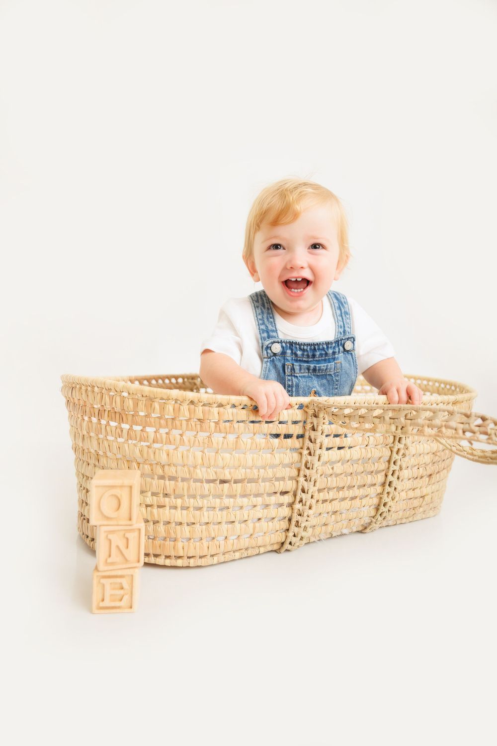 Happy baby in denim overalls sitting in a wicker Moses basket with wooden ONE blocks, celebrating first birthday.