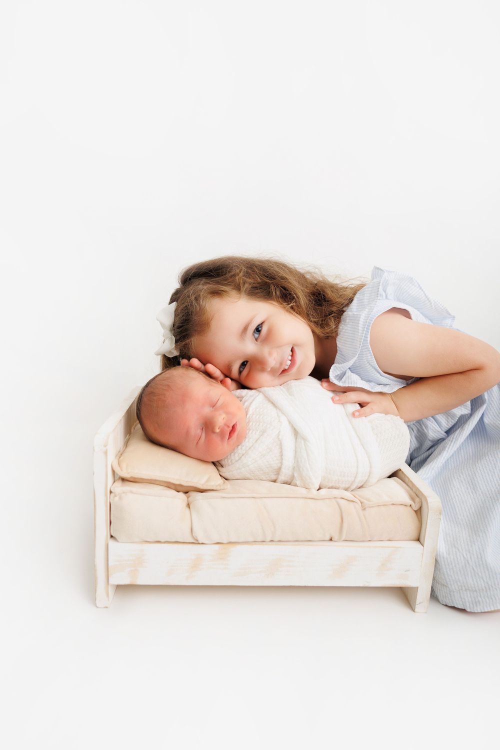 A smiling young girl in a blue dress poses with her swaddled newborn sibling lying in a small wooden bed.