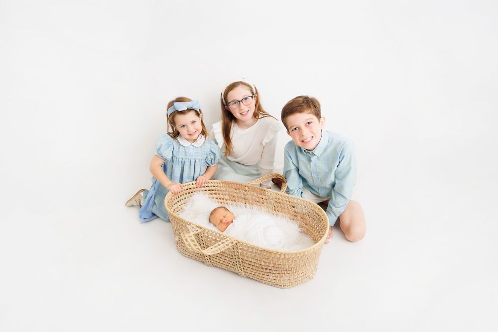 Three siblings pose with newborn baby in wicker bassinet on white background in professional studio photo.