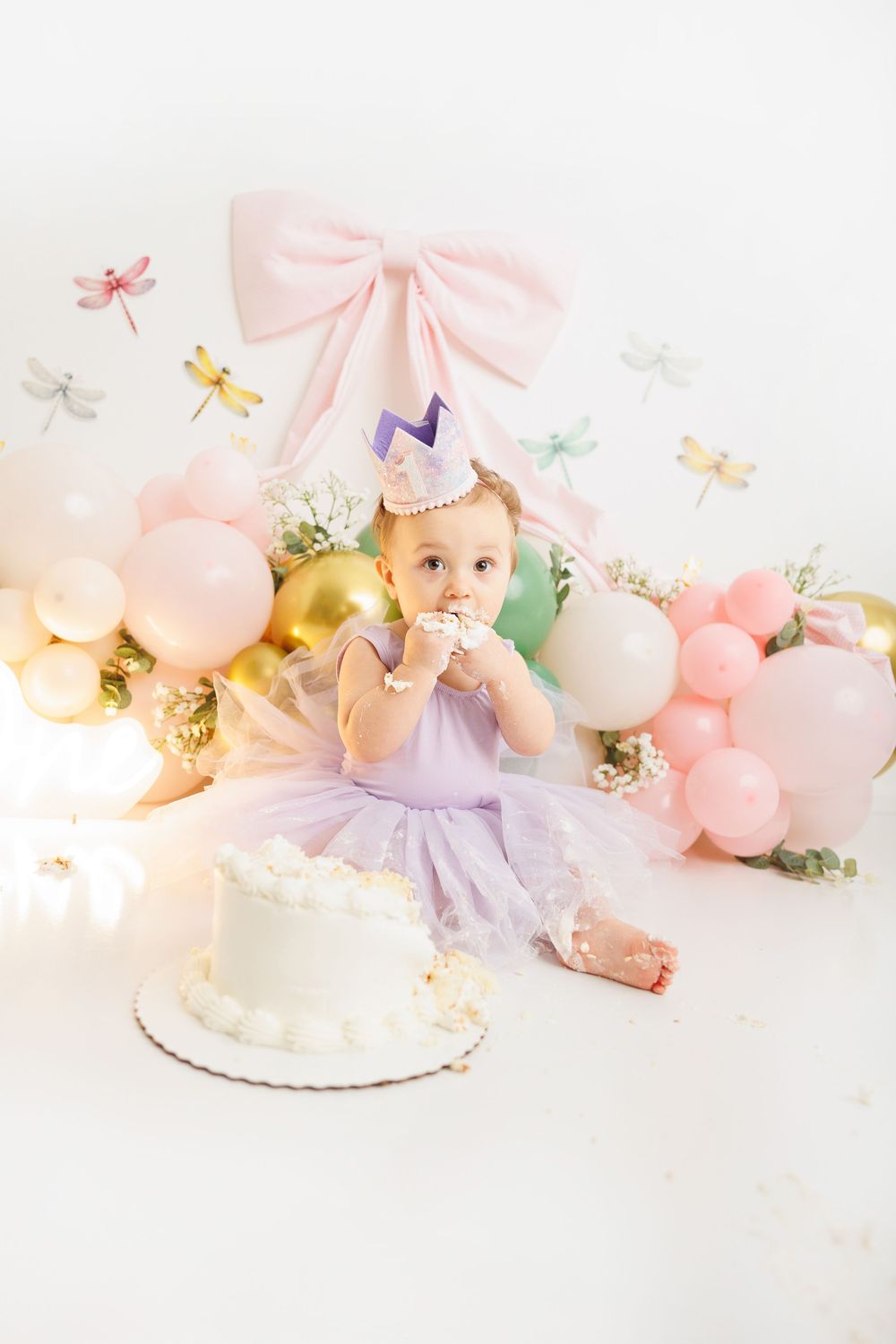 Baby girl in purple tutu enjoys first birthday cake smash with pastel balloons and dragonfly decorations.
