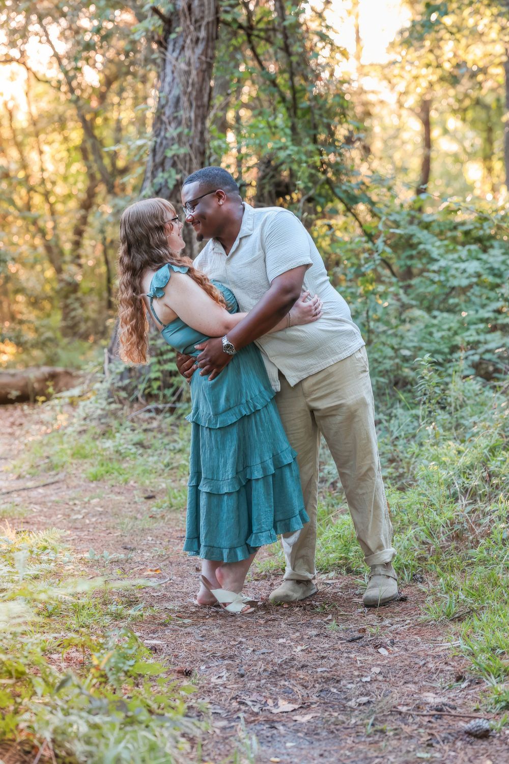 A couple embraces on a sunlit forest path while wearing casual summer outfits during a romantic afternoon.