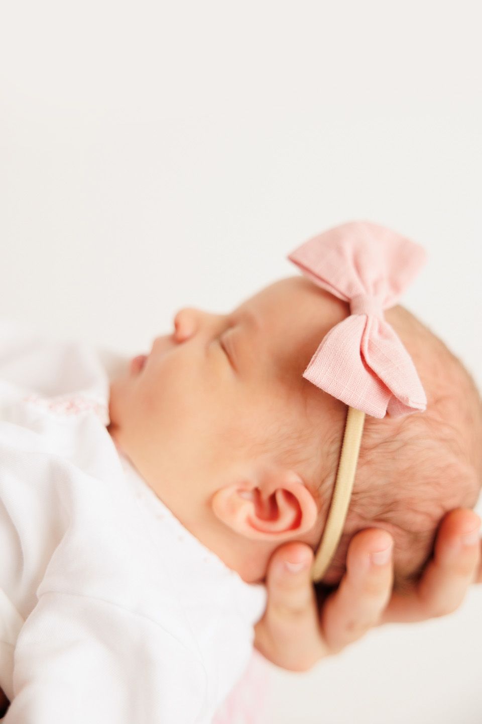 Sleeping newborn baby girl wearing a pink bow headband and white outfit, peaceful close-up portrait.