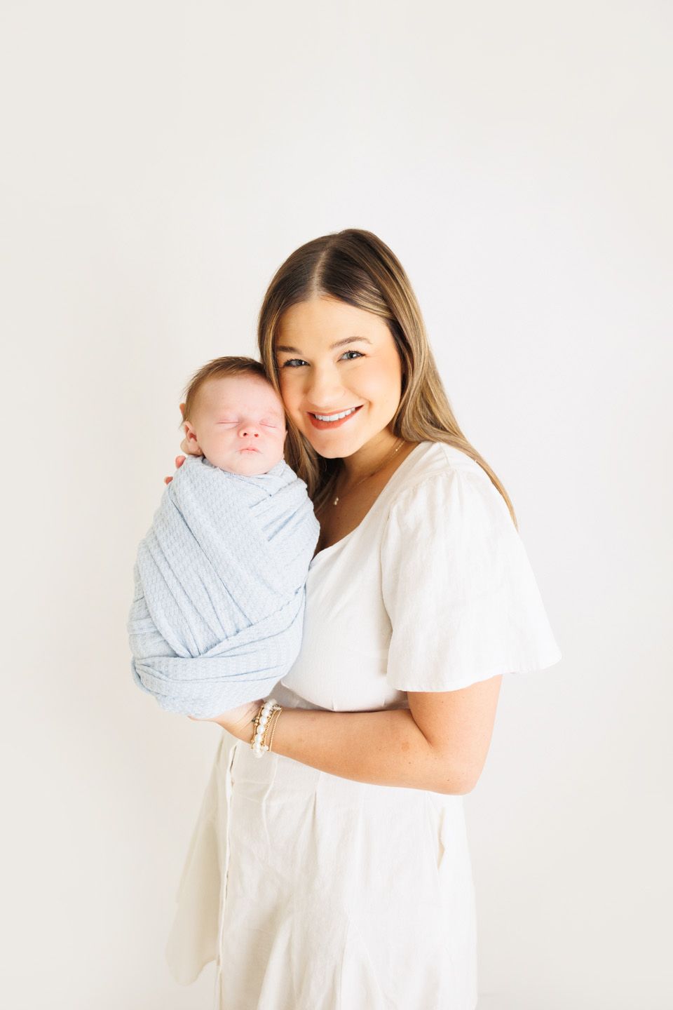 Smiling mother in white dress holding swaddled newborn baby in light blue wrap against white background.
