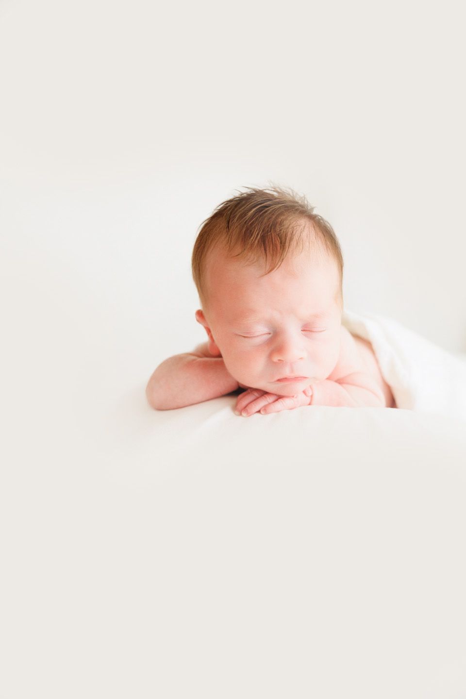 Peaceful newborn baby sleeping with hands tucked under chin on soft white blanket.