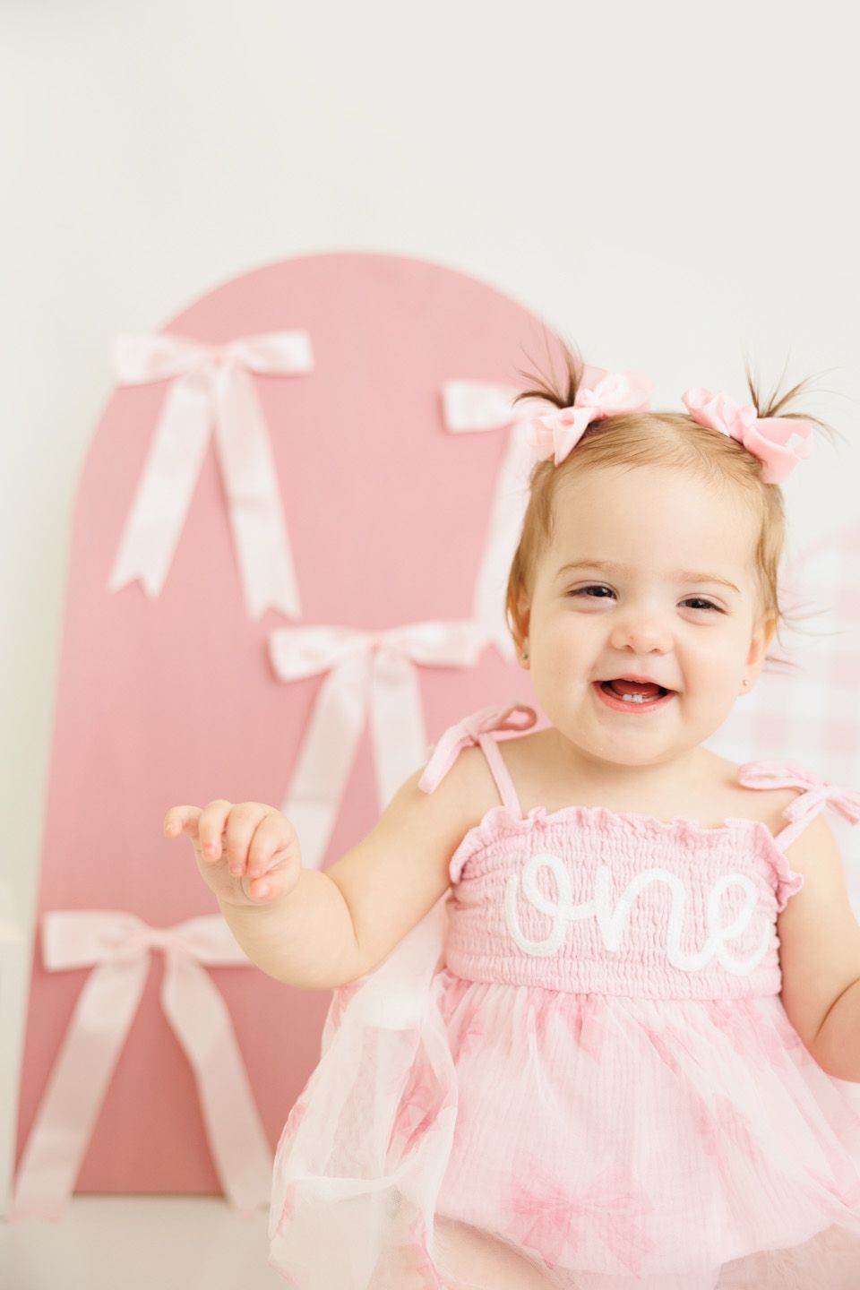 Smiling baby girl in pink dress with 'one' embroidery celebrates first birthday in front of pink bow backdrop.