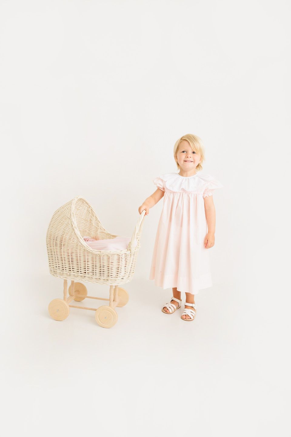 Young girl in pink smocked dress with white collar stands beside a wicker doll pram on white background.