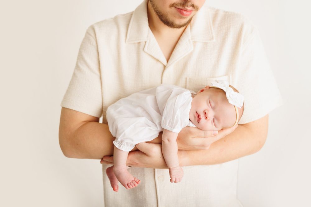 A man in a cream shirt gently cradles a sleeping newborn baby dressed in white against a light background.