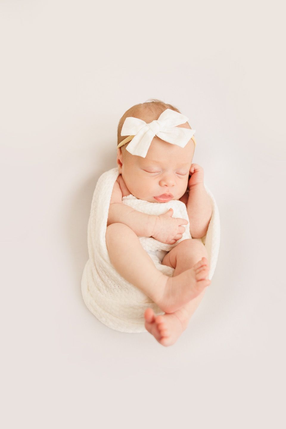 Sleeping newborn baby girl wrapped in white swaddle with bow headband on white background.