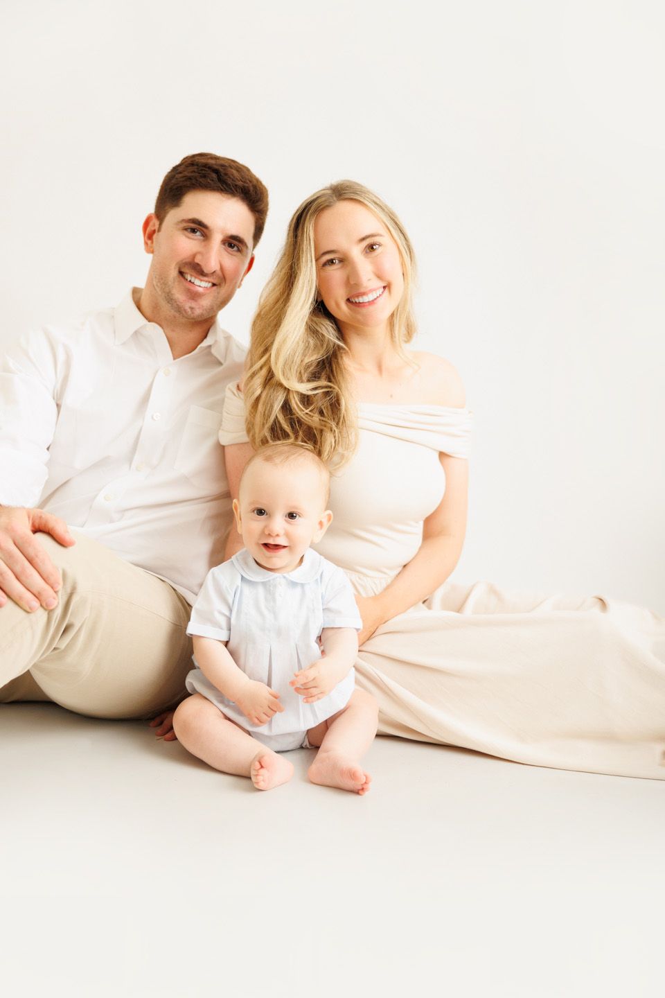 A smiling family with a man, woman, and baby sitting together on a white background in neutral tones.
