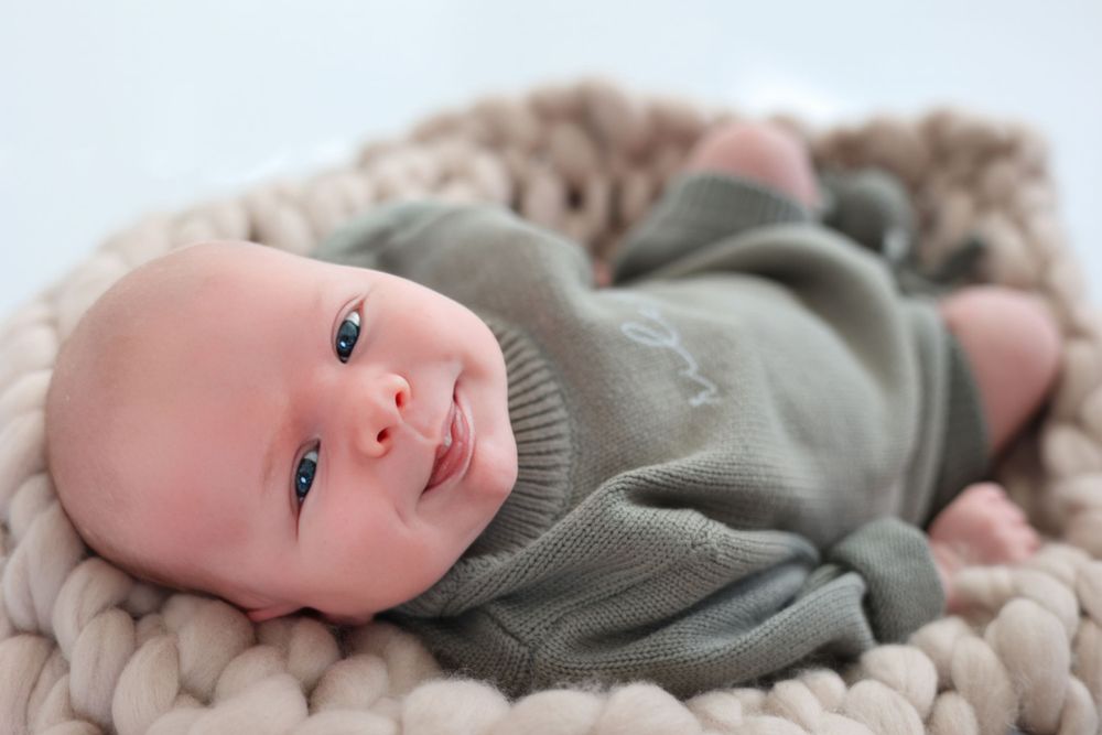 Baby in gray sweater lying on chunky knit blanket looking at camera. Newborn baby photography session.