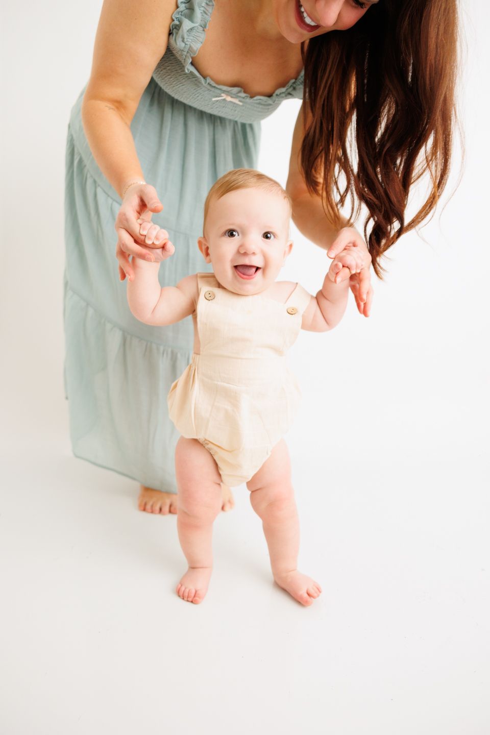 Happy baby in cream romper takes first steps with smiling brunette mother in mint dress on white background.