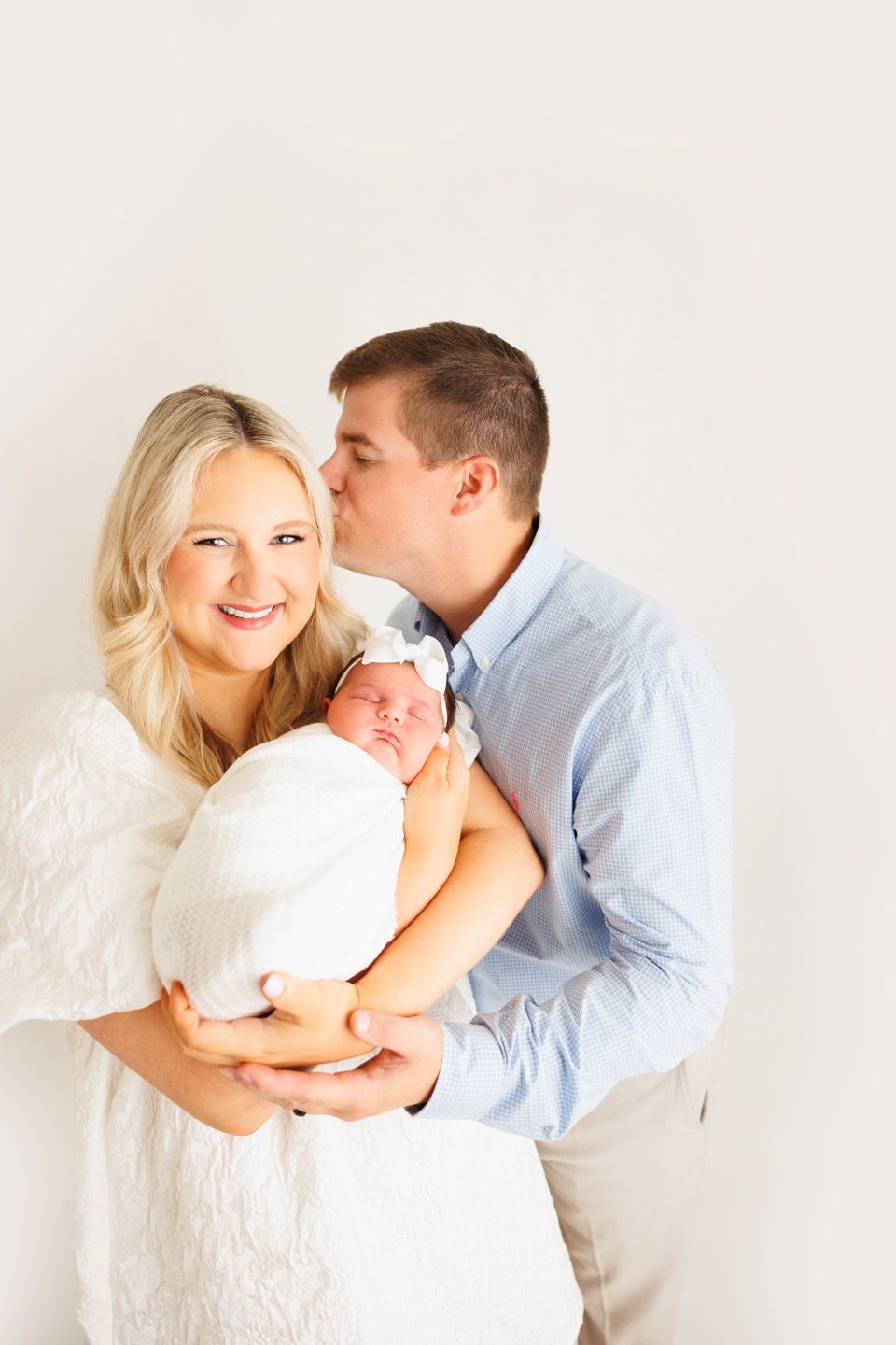 A couple in a bright studio holding their swaddled newborn baby, the man kissing the smiling woman on the cheek.