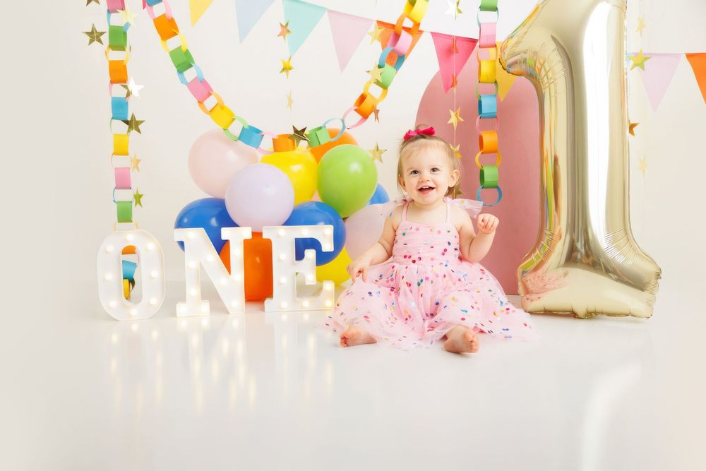 Baby girl in pink dress celebrates first birthday with colorful balloons, ONE sign, and gold number 1 balloon.