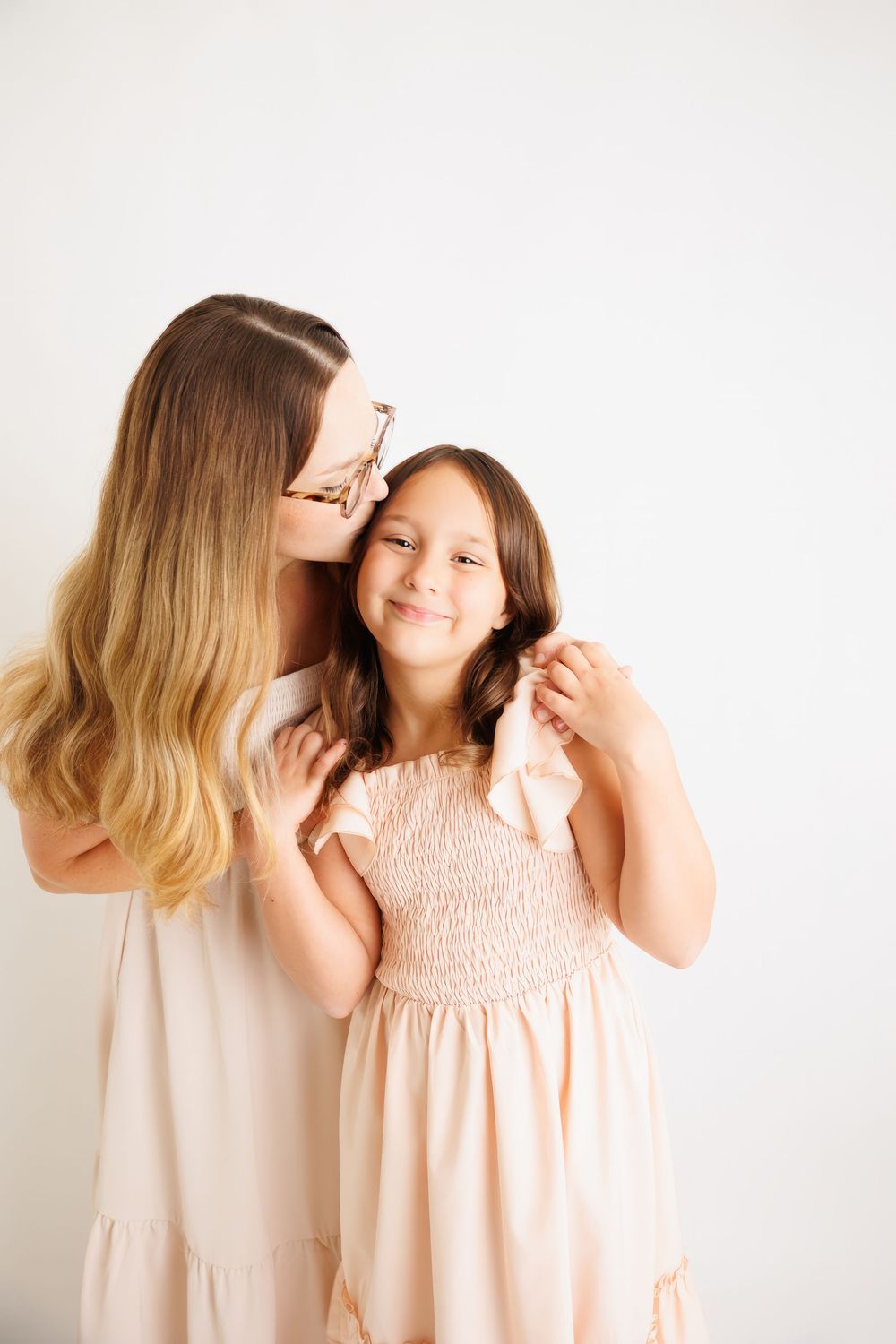 A woman in a pink dress kisses a young girl in a matching dress, both smiling warmly against a white background.