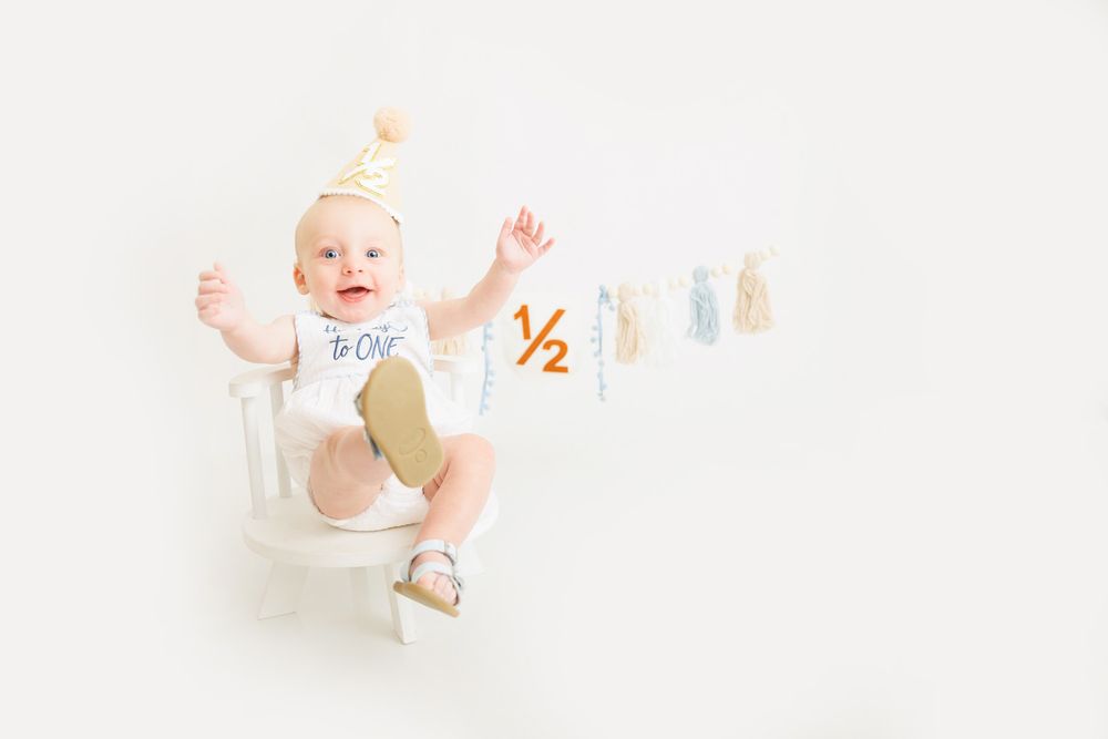 Baby in white onesie and gold party hat sits on chair celebrating half birthday with decorative 1/2 banner.