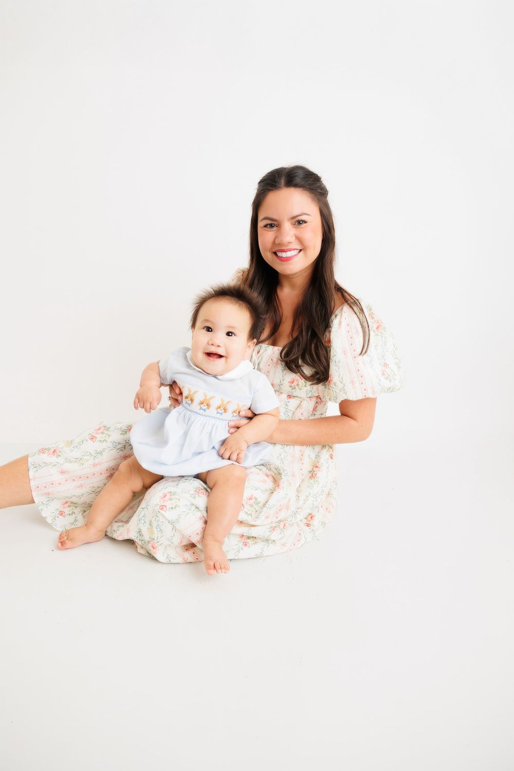 Smiling mother in floral dress sitting with happy baby in white outfit on white background.