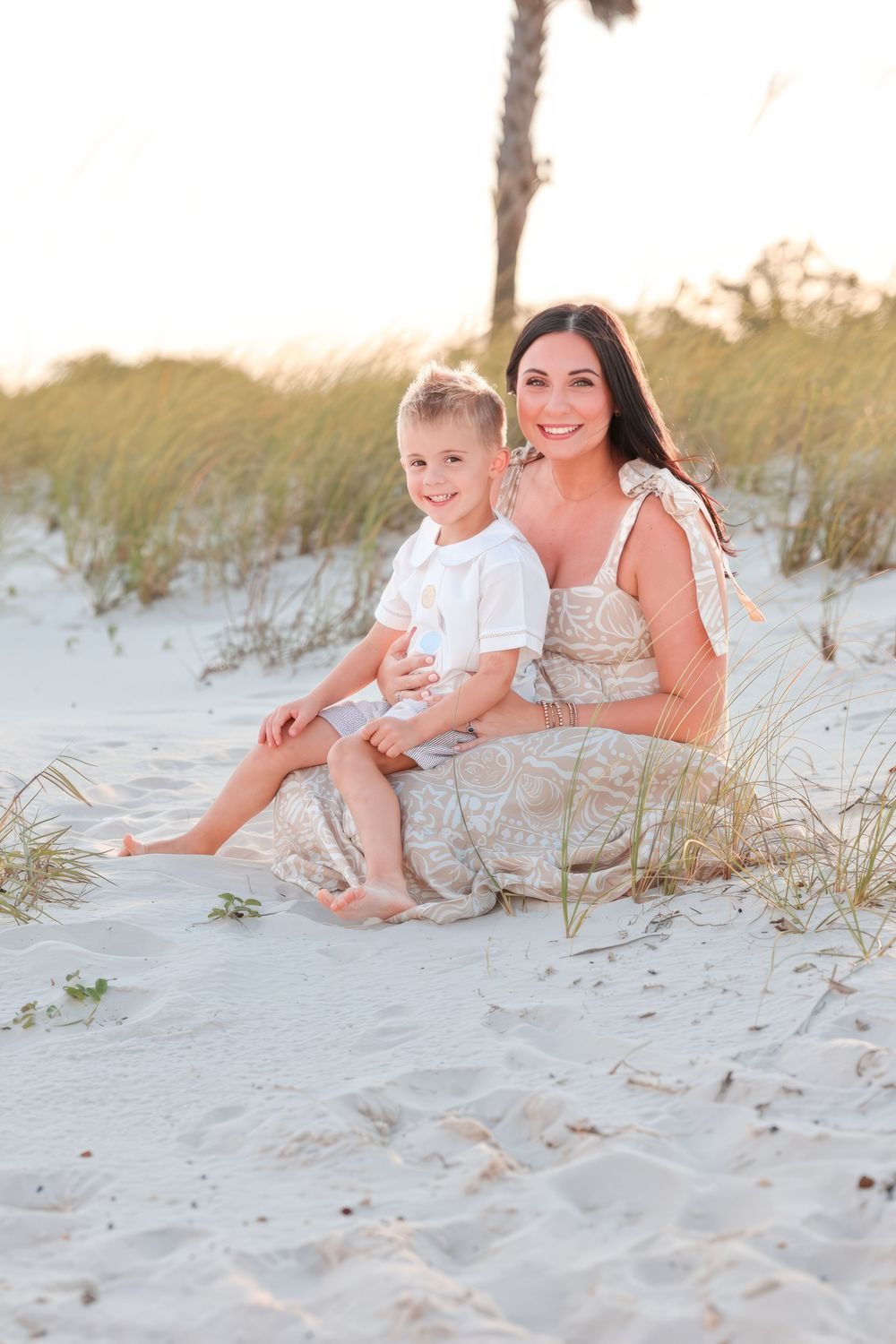 Mother and child sit together on white sandy beach dunes during golden sunset hour for family photos.