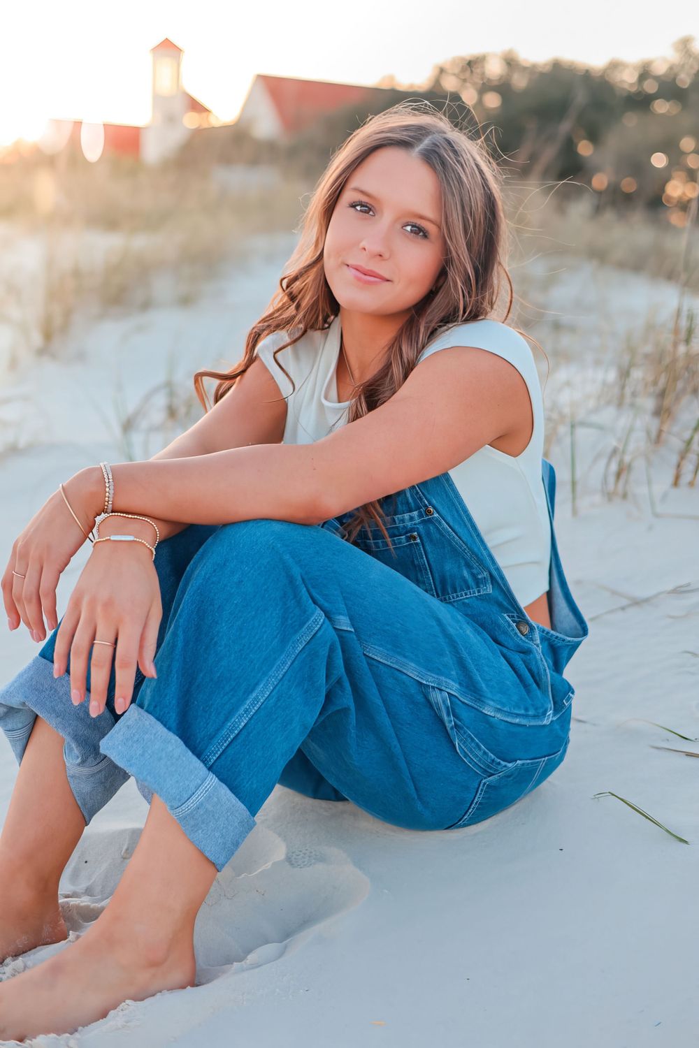 Young woman in overalls sitting on beach sand during golden hour. Senior high school graduation casuals.