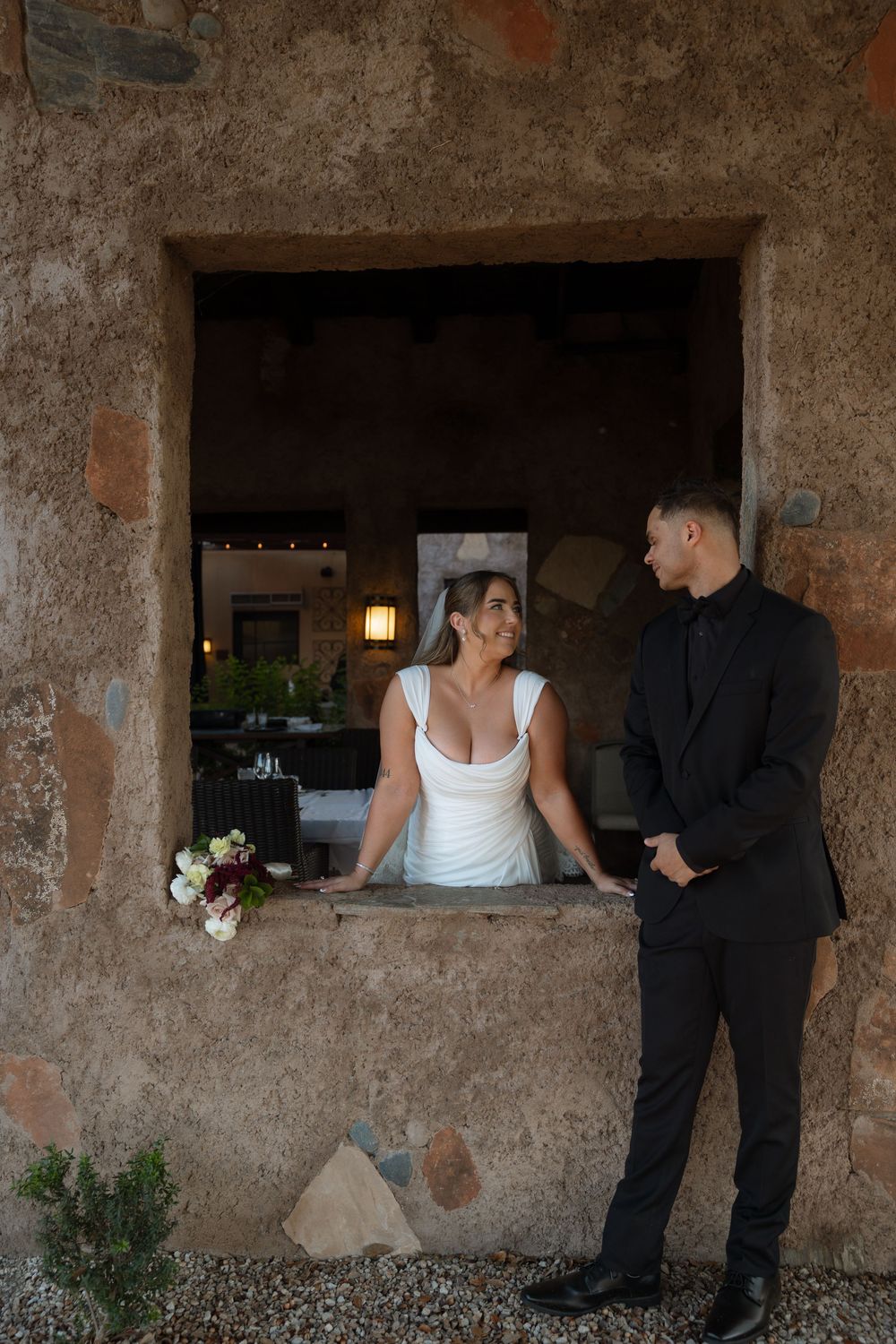 Bride in white gown and groom in black suit share a romantic moment by a rustic stone window at their wedding venue.