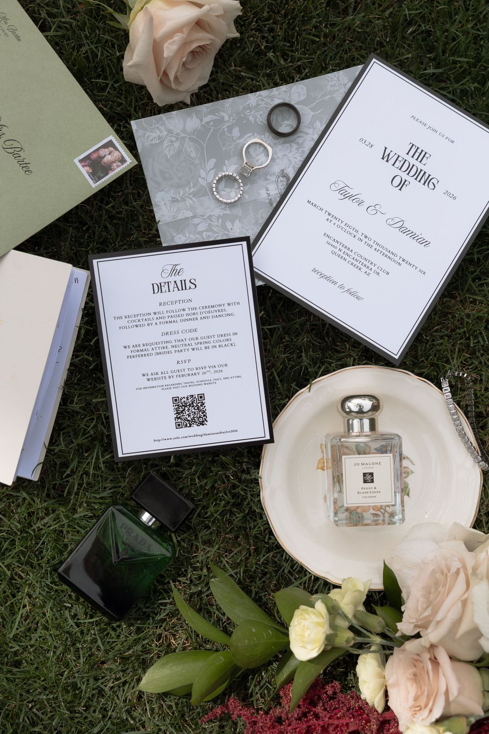 Elegant wedding flat lay with invitation suite, Jo Malone perfume, rings, and blush roses on green grass.