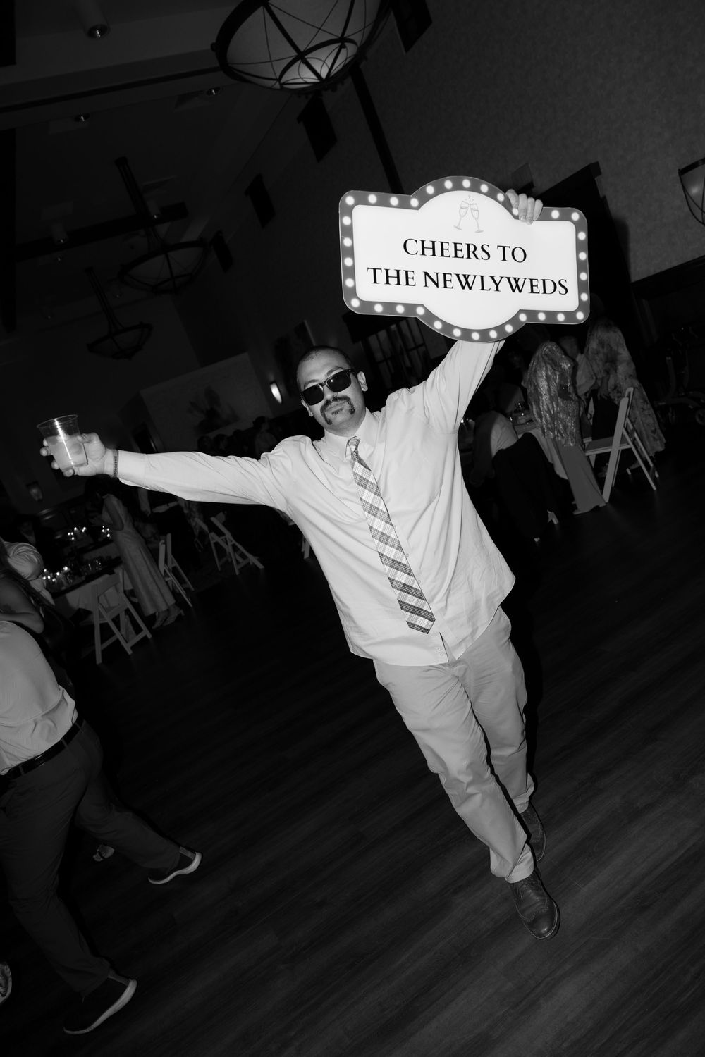 Man in sunglasses holding a lit Cheers to the Newlyweds sign at a wedding reception celebration.