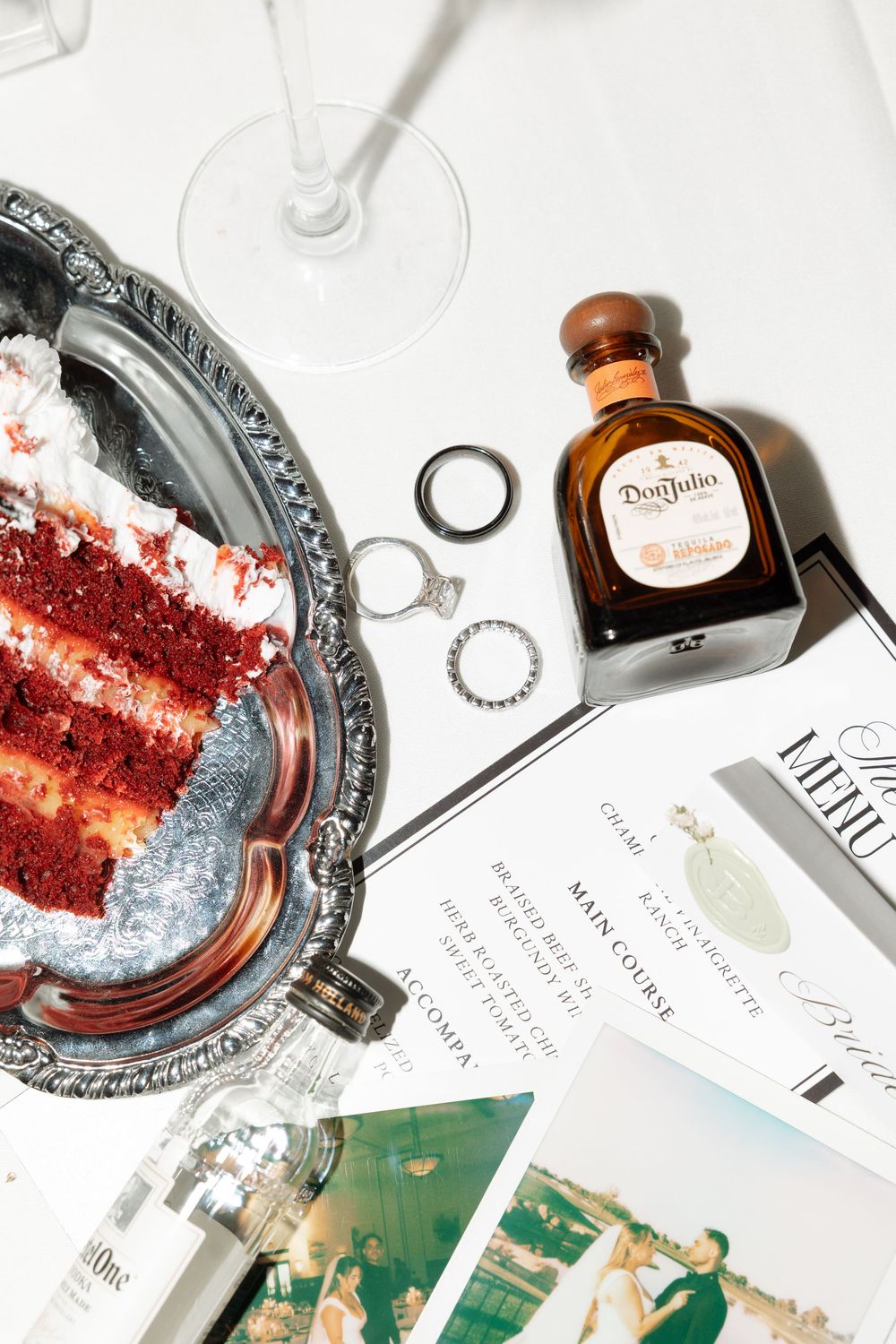 Red velvet cake slice, Don Julio tequila, wedding rings, menu card, and Polaroid photos on a white table.