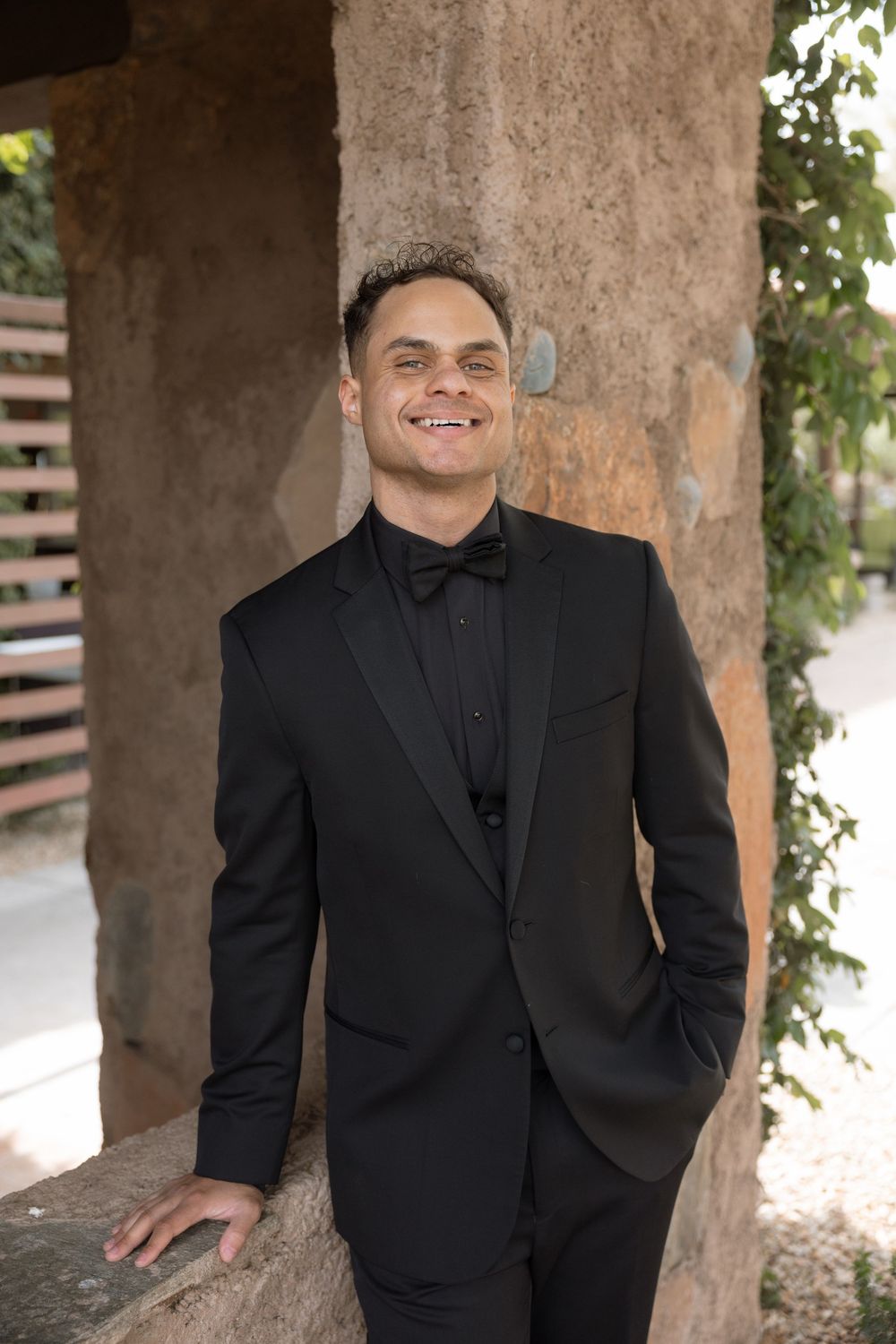 Smiling groom in black tuxedo with bow tie leans against a stone pillar in an outdoor wedding venue setting.