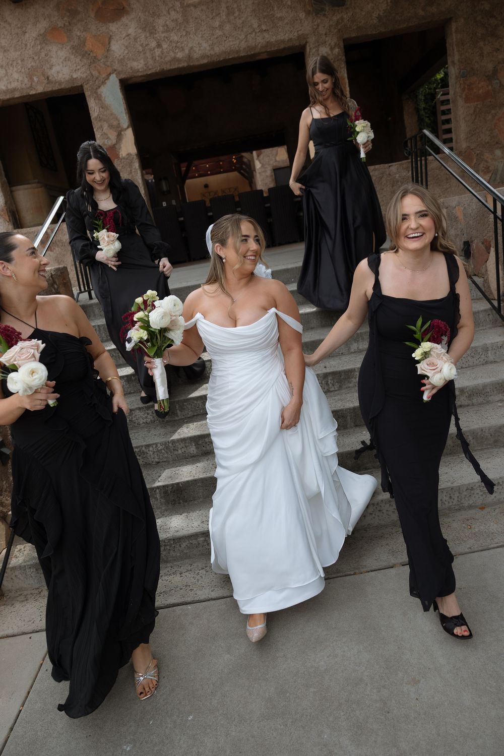 Bride in white off-shoulder gown walks with bridesmaids in black dresses carrying bouquets down stone steps.