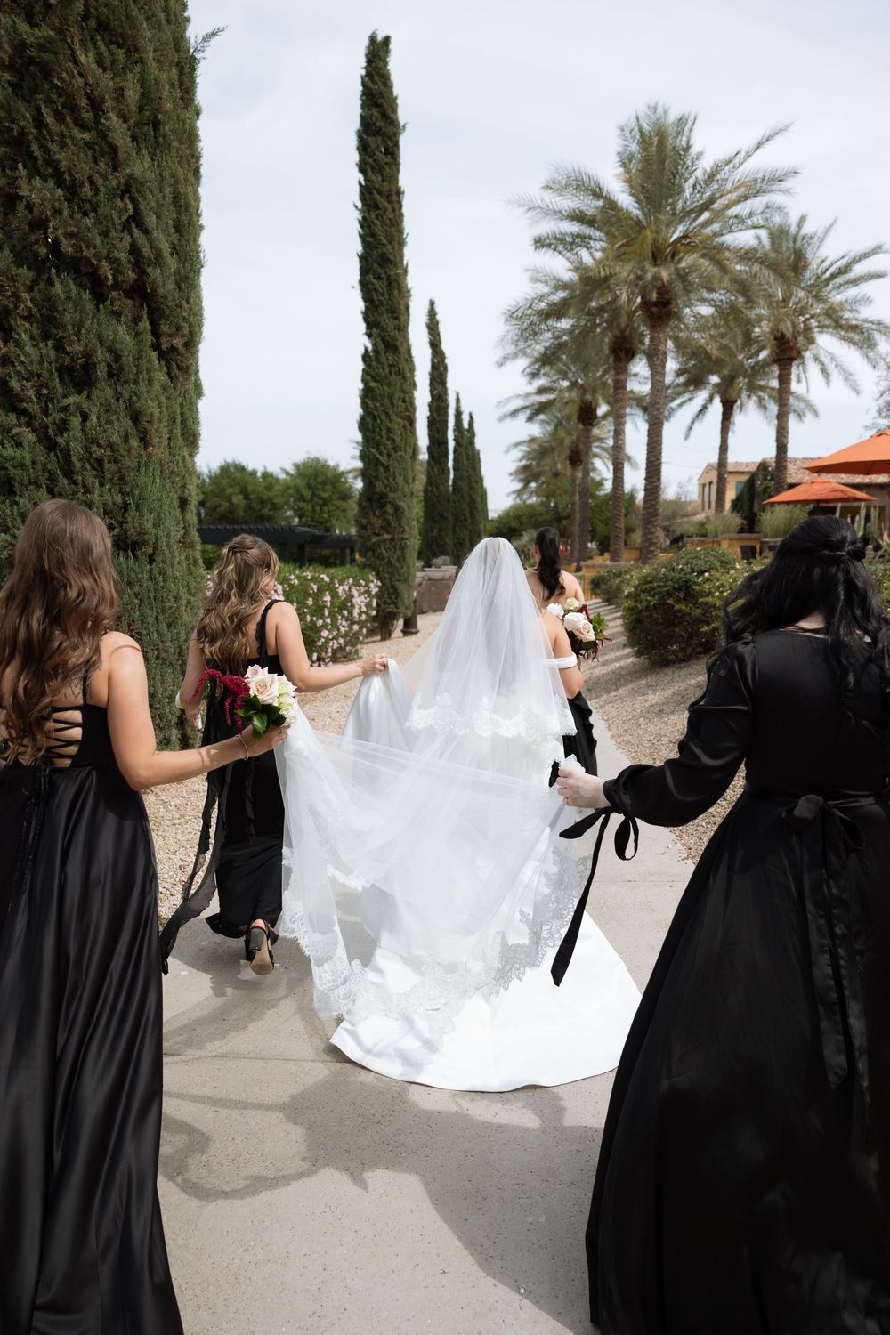Bride in white gown and veil walks with bridesmaids in black dresses along a garden path lined with cypress and palm trees.