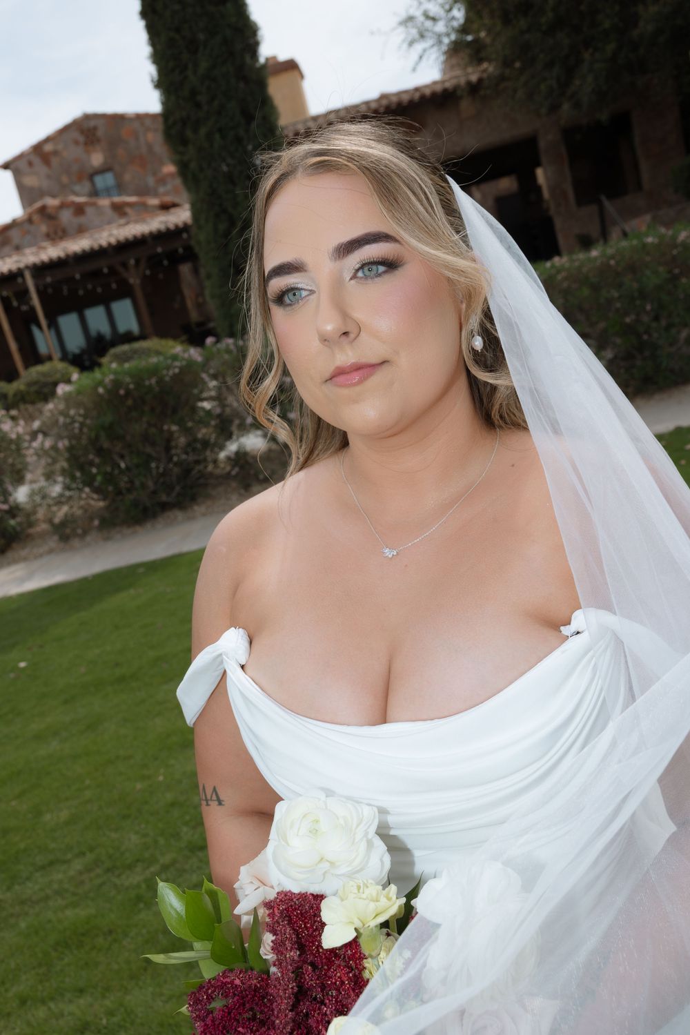 Bride in white off-shoulder gown and veil holding bouquet of white and burgundy flowers outdoors.