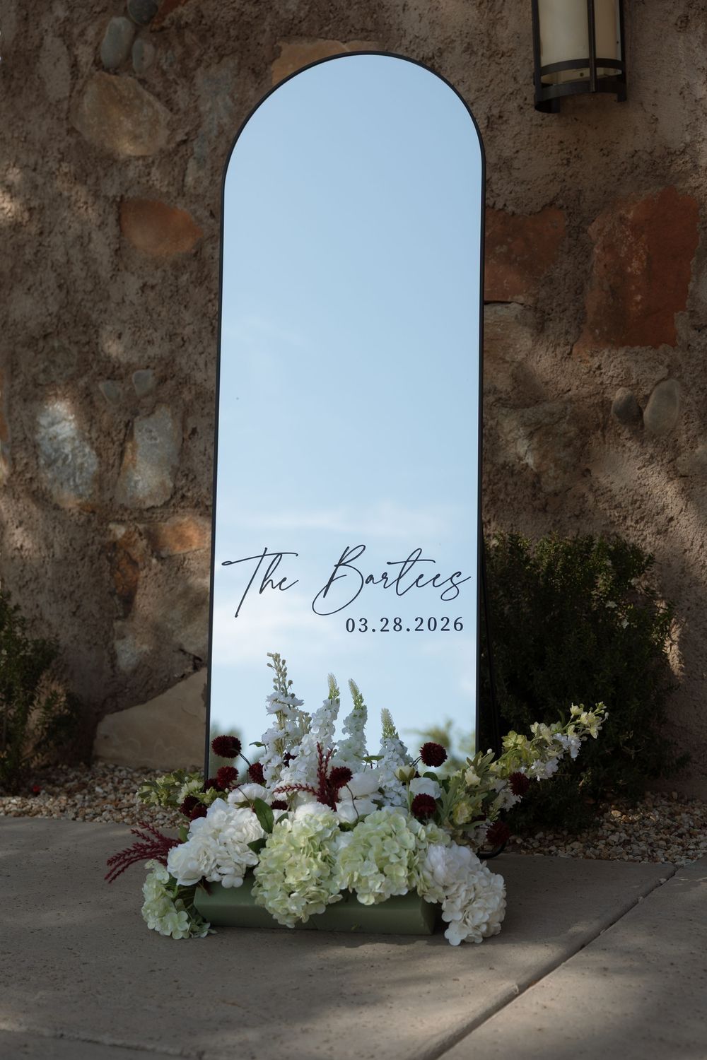 Elegant arched mirror with The Bartlees 03.28.2026 wedding sign, adorned with white and burgundy floral arrangement outdoors.