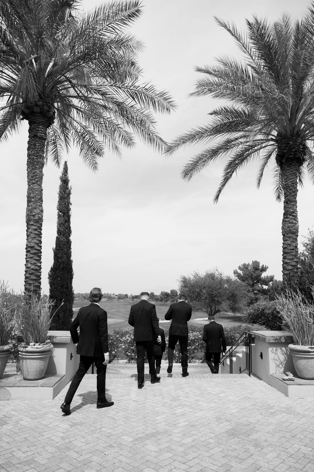 Black and white photo of four men in suits walking between tall palm trees on a brick patio.