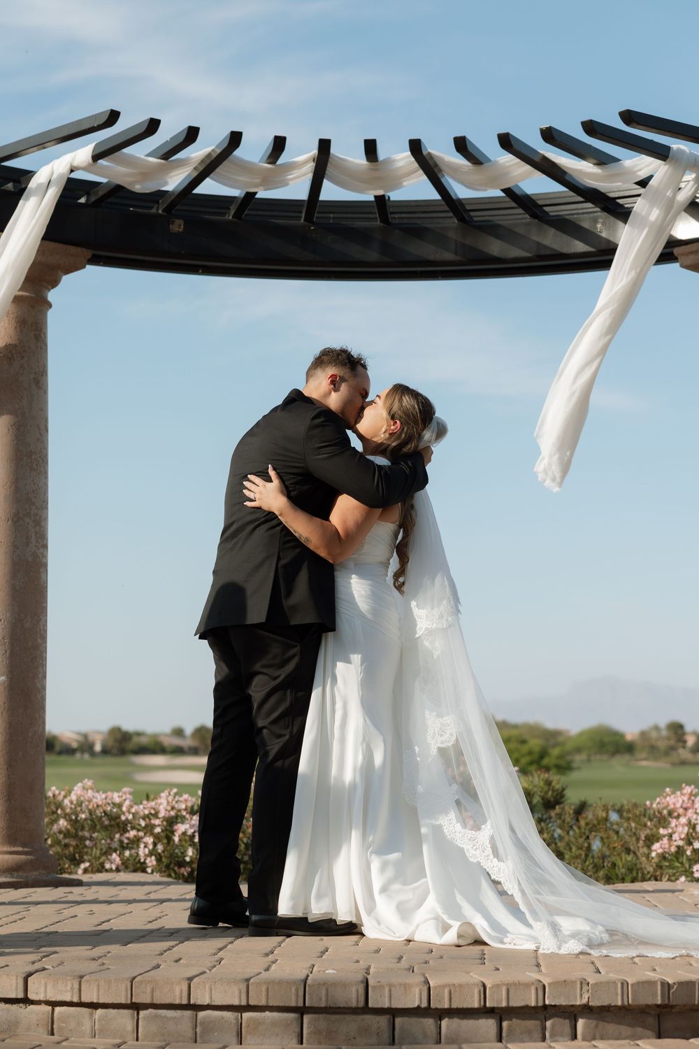 Bride and groom share first kiss under elegant pergola with scenic green landscape and blue sky backdrop.
